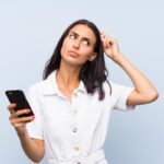 Young woman with a mobile phone over isolated blue wall having doubts and with confuse face expression